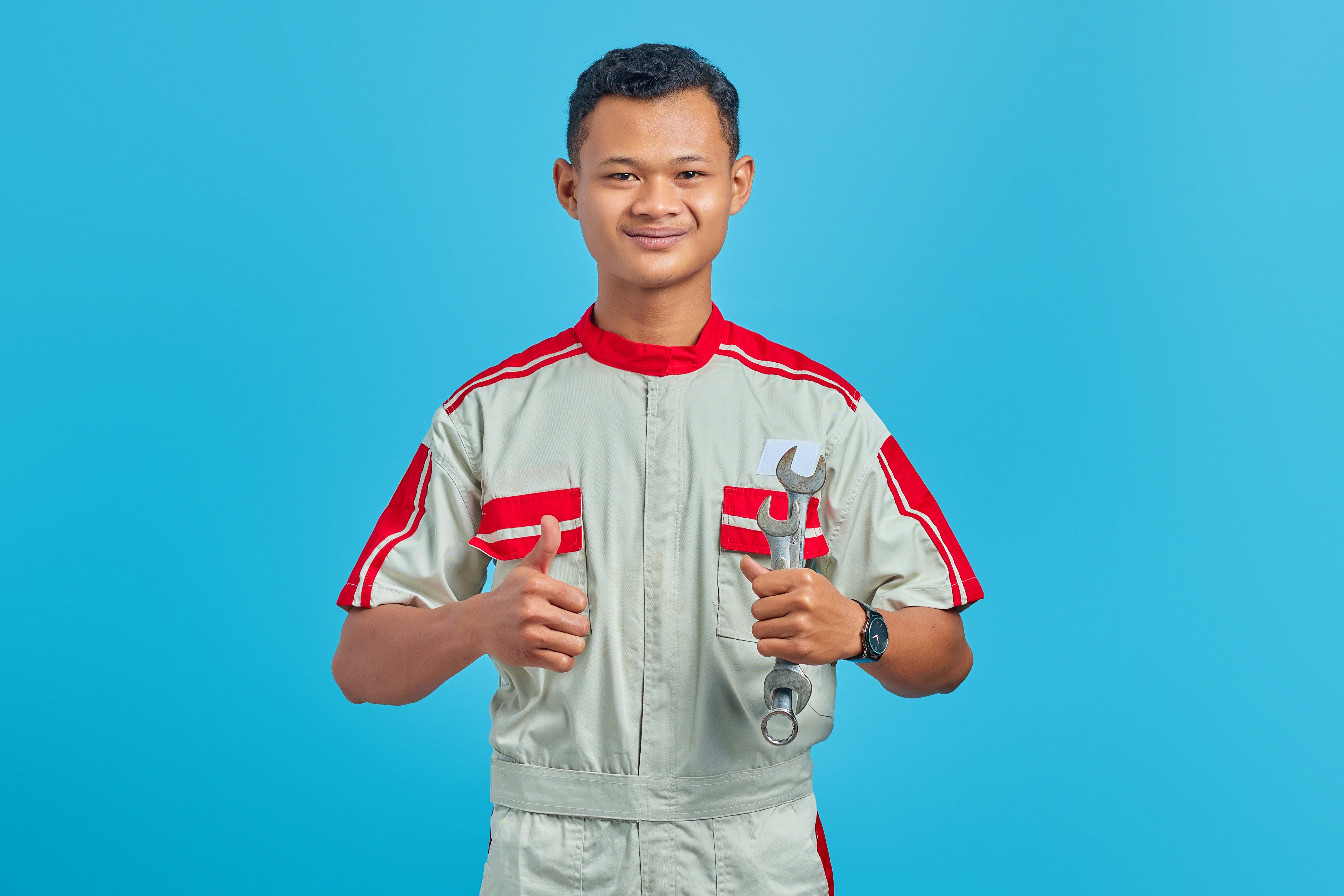 Portrait of smiling young Asian mechanic holding wrench and showing thumbs up to camera isolated on blue background